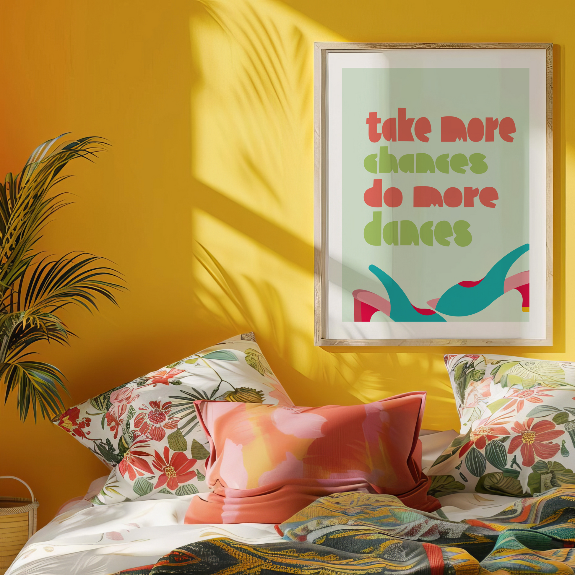 A framed motivational wall art print with "Take More Chances, Do More Dances" in bold colours, displayed in a vibrant bedroom with floral cushions and a yellow wall.