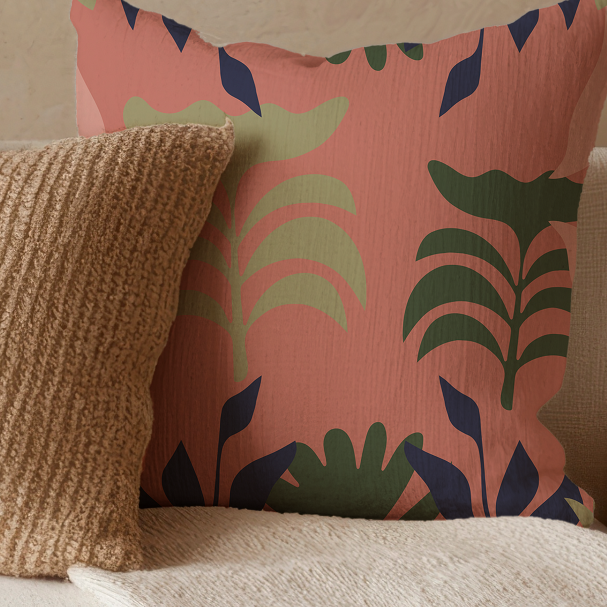 A square decorative throw pillow with a terracotta pink background and a botanical leaf pattern in dark green and navy blue, styled on a textured cream sofa next to a chunky beige knit pillow.