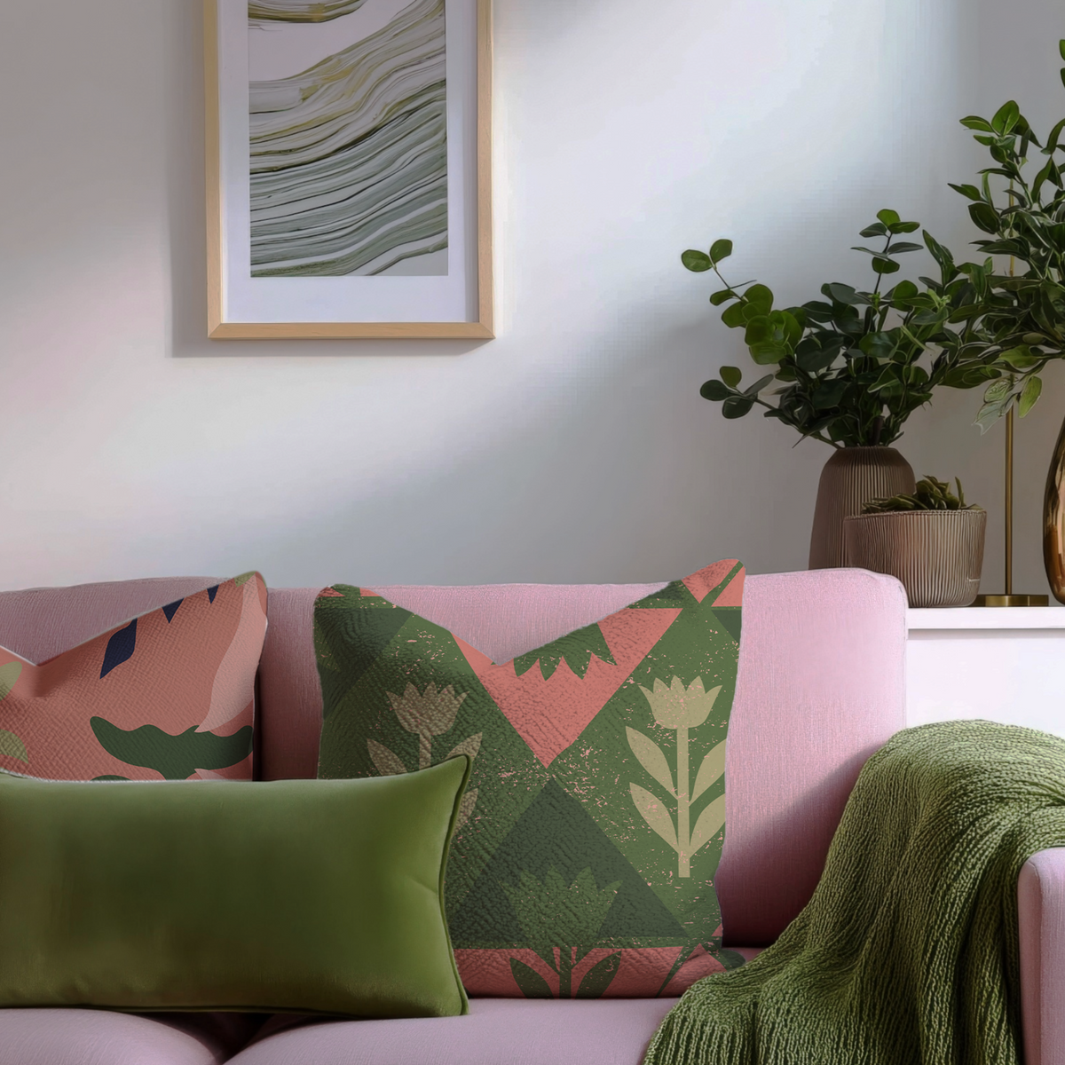 Three decorative throw pillows arranged on a pink sofa in a styled living room setting, with a green knit throw blanket draped over the arm and a framed abstract print on the wall.