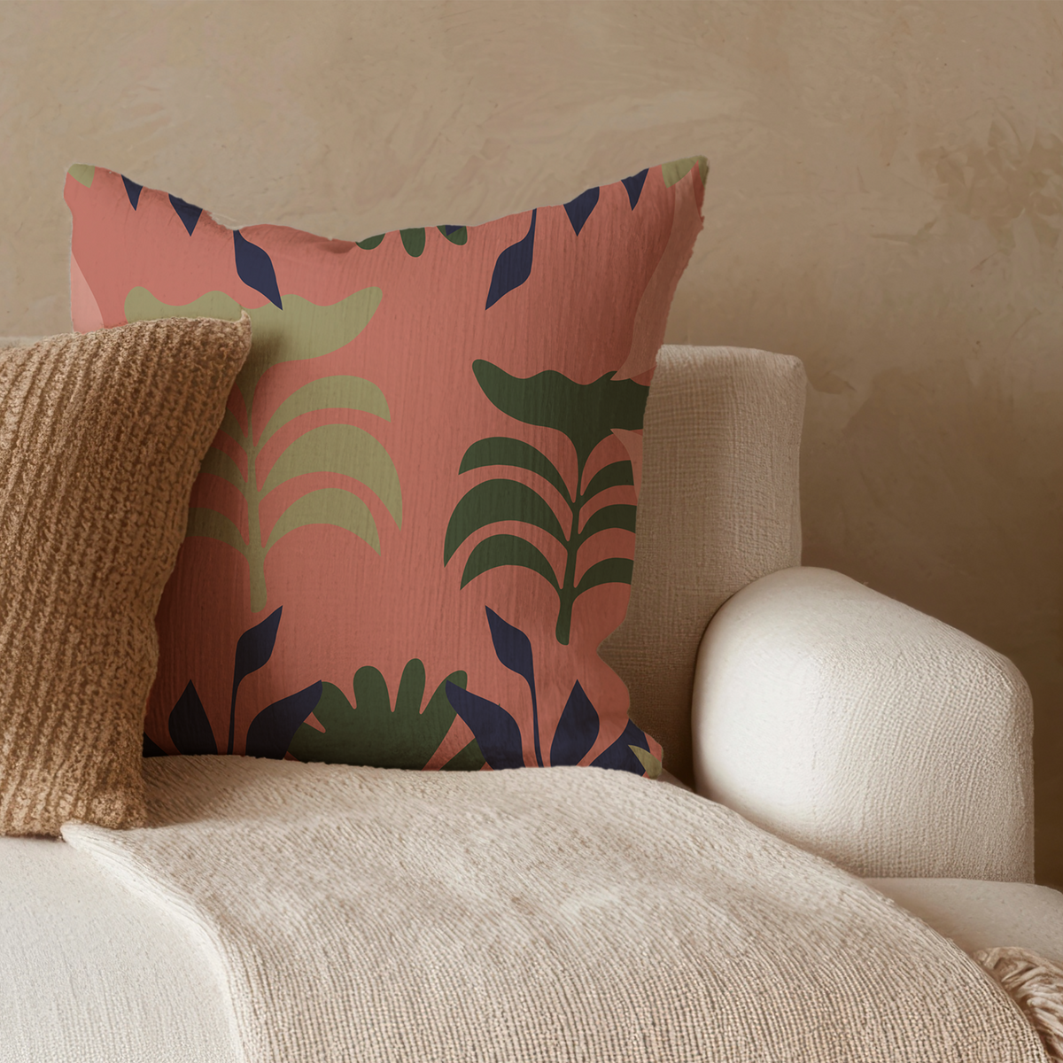A square decorative throw pillow with a terracotta pink background and a green, dark blue, and sage botanical pattern, styled on a cream-colored armchair next to a textured brown knit pillow.