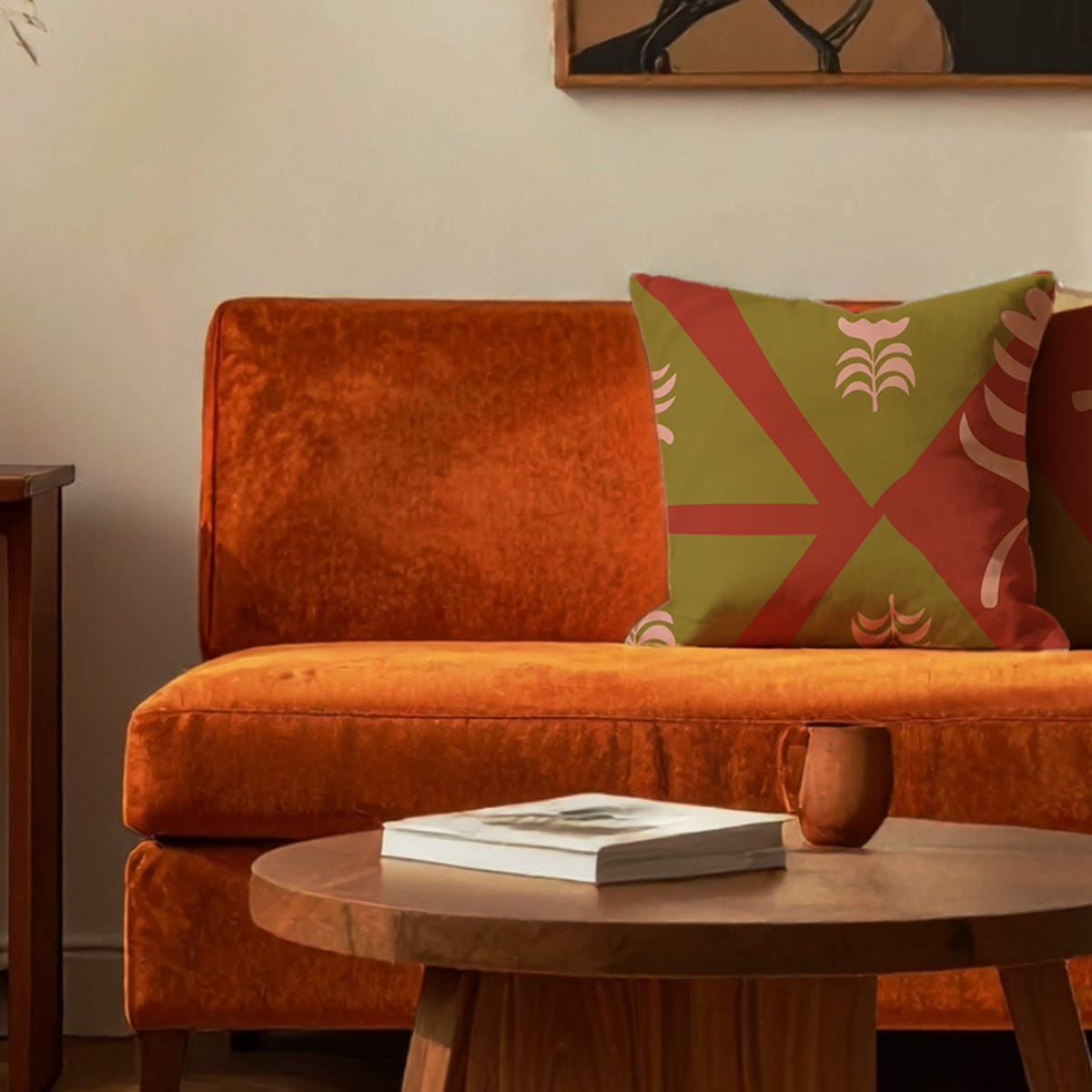 An orange velvet armchair in a styled living room setting, featuring a green and red patterned accent pillow, a round wooden coffee table with a book and mug, and a cream-colored wall.