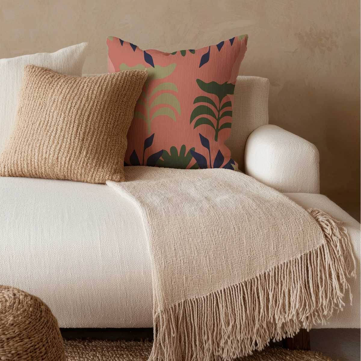 A styled living room scene showing a cream fringed throw blanket draped over a white armchair, with two decorative pillows including a coral pink one with a green and navy botanical pattern.