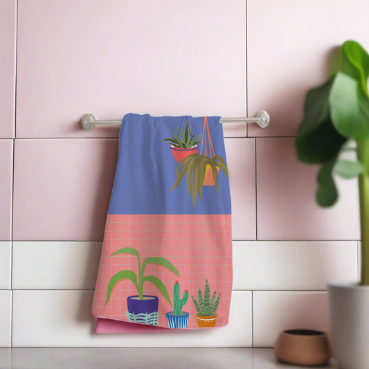 a pink and blue colour block kitchen towel with a grid design and house plants on the front of it 